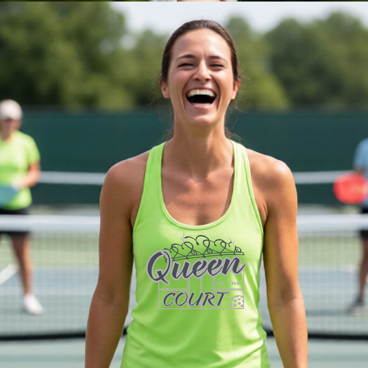 A woman laughing on a pickleball court, wearing a lime green racerback tank top with a graphic that reads 'Queen of the Court' in gray and white and a pickleball graphic. 