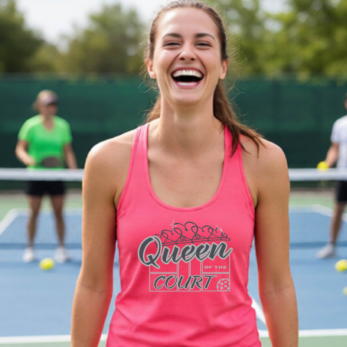 A woman laughing on a pickleball court, wearing a coral pink racerback tank top with a graphic that reads 'Queen of the Court' in gray and white and a pickleball graphic. 
