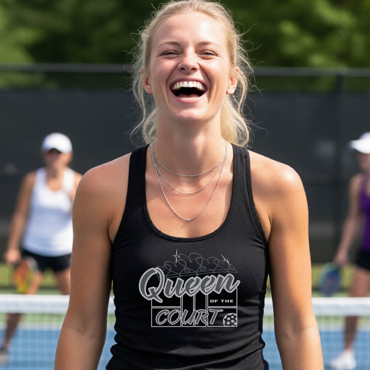 A woman laughing on a pickleball court, wearing a black racerback tank top with a graphic that reads 'Queen of the Court' in gray and white and a pickleball graphic. 