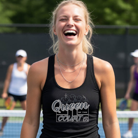 A woman laughing on a pickleball court, wearing a black racerback tank top with a graphic that reads 'Queen of the Court' in gray and white and a pickleball graphic. 