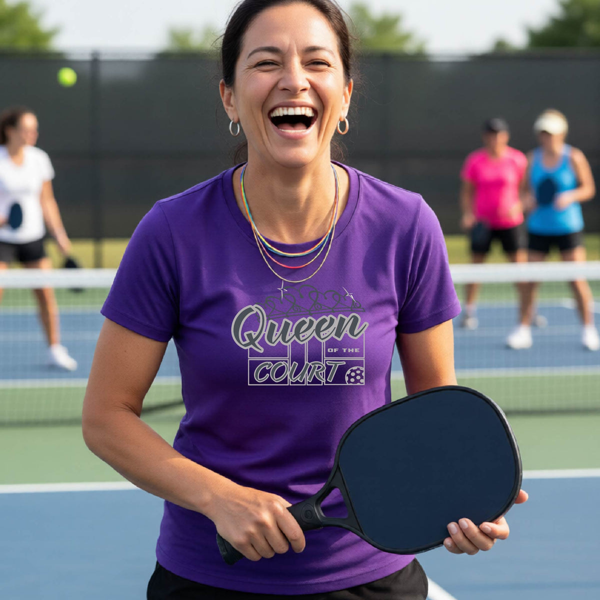 A woman laughing on a pickleball court wearing a purple t-shirt with a blue and white graphic that reads 'Queen COURT' with a pickleball and court motiff, standing on a pickleball court. 