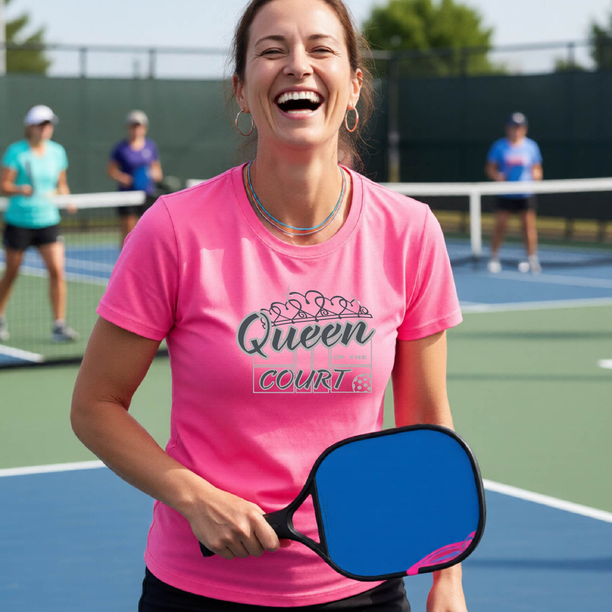 A woman laughing on a pickleball court wearing a pink t-shirt with a blue and white graphic that reads 'Queen COURT' with a pickleball and court motiff, standing on a pickleball court. 