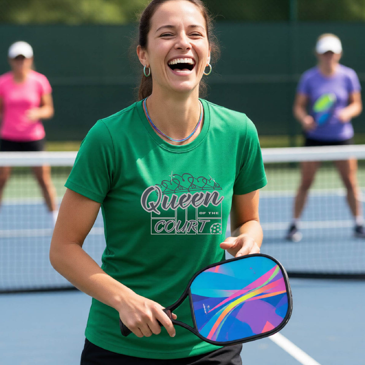 A woman laughing on a pickleball court wearing a green t-shirt with a blue and white graphic that reads 'Queen COURT' with a pickleball and court motiff, standing on a pickleball court. 