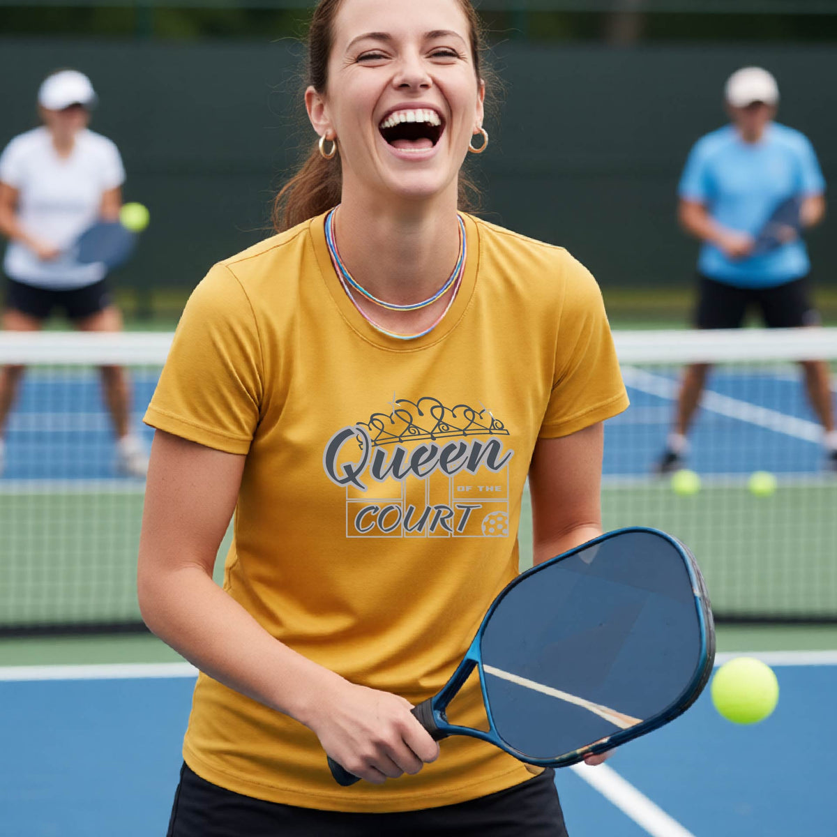A woman laughing on a pickleball court wearing a mustard yellow t-shirt with a blue and white graphic that reads 'Queen COURT' with a pickleball and court motiff, standing on a pickleball court. 