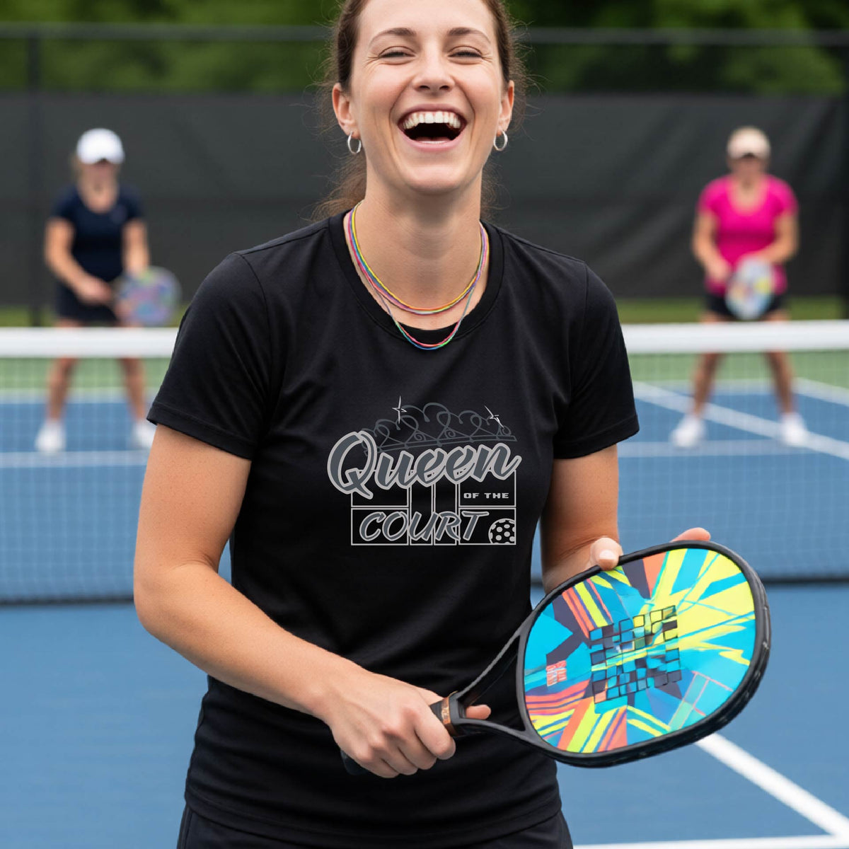 A woman laughing on a pickleball court wearing a black t-shirt with a blue and white graphic that reads 'Queen COURT' with a pickleball and court motiff, standing on a pickleball court. 