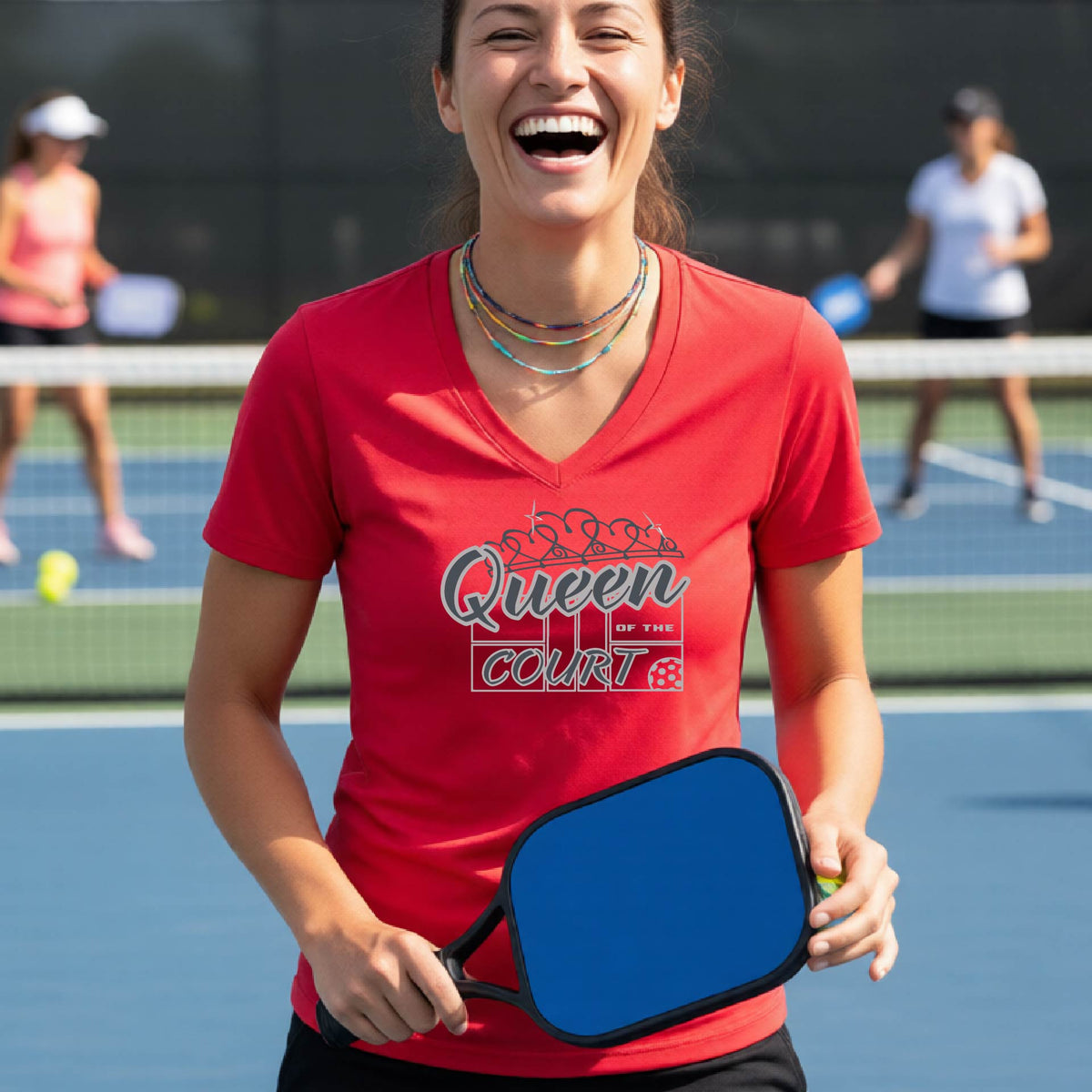 A woman laughing on a pickleball court wearing a red v-neck t-shirt with a white graphic that reads 'Queen of the Court.