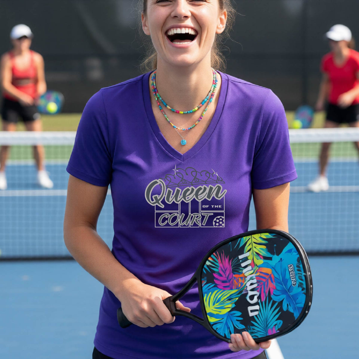 A woman laughing on a pickleball court wearing a purple v-neck t-shirt with a white graphic that reads 'Queen of the Court.