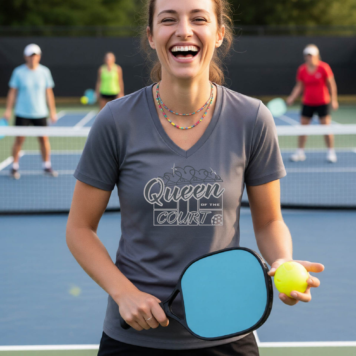 A woman laughing on a pickleball court wearing a gray v-neck t-shirt with a white graphic that reads 'Queen of the Court.