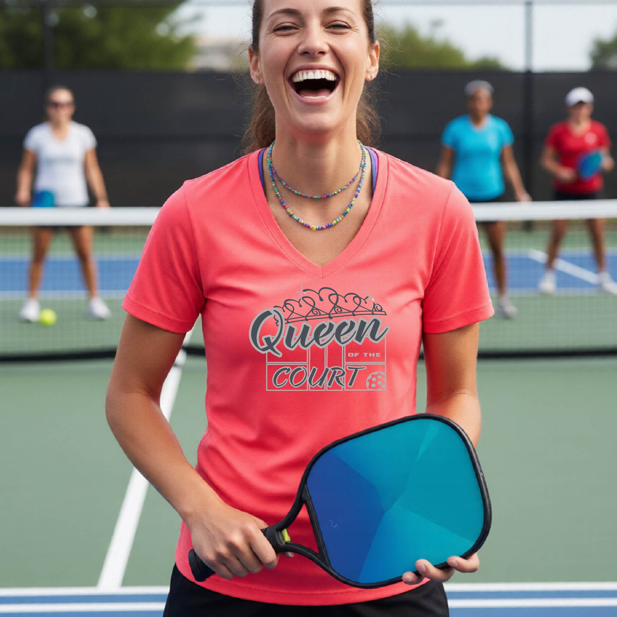 A woman laughing on a pickleball court wearing a coral pink v-neck t-shirt with a white graphic that reads 'Queen of the Court.