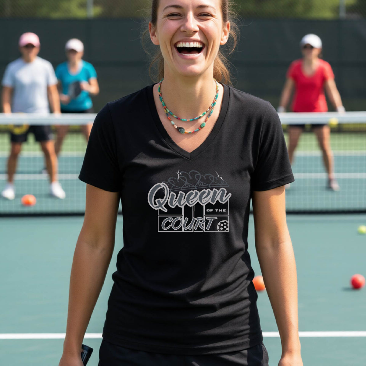 A woman laughing on a pickleball court wearing a black v-neck t-shirt with a white graphic that reads 'Queen of the Court.