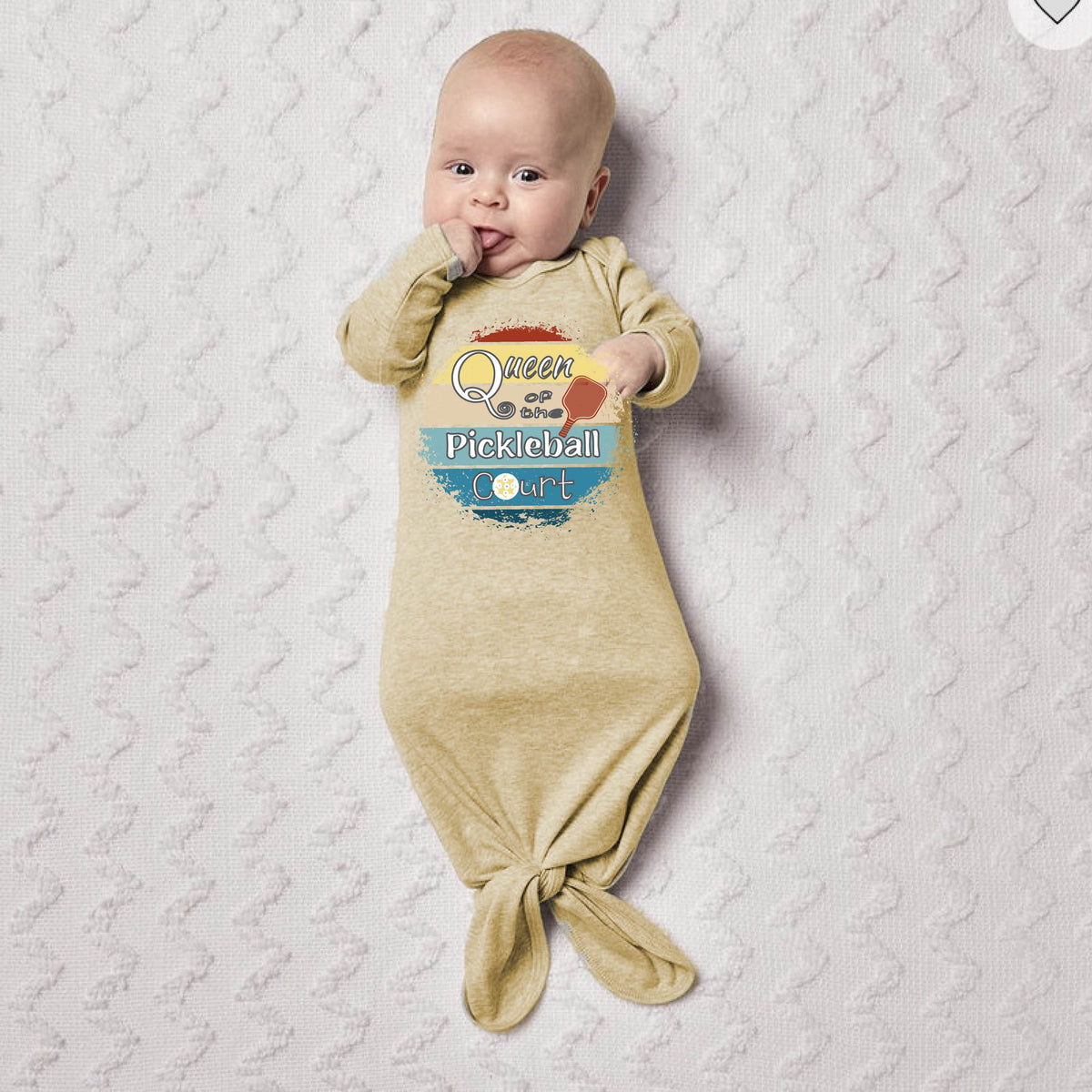 A baby a blanket, wearing a beige footless onesie with a colorful 'Queen of hte Pickleball Court' graphic. A chain-link fence and pickleball court are visible in the background.