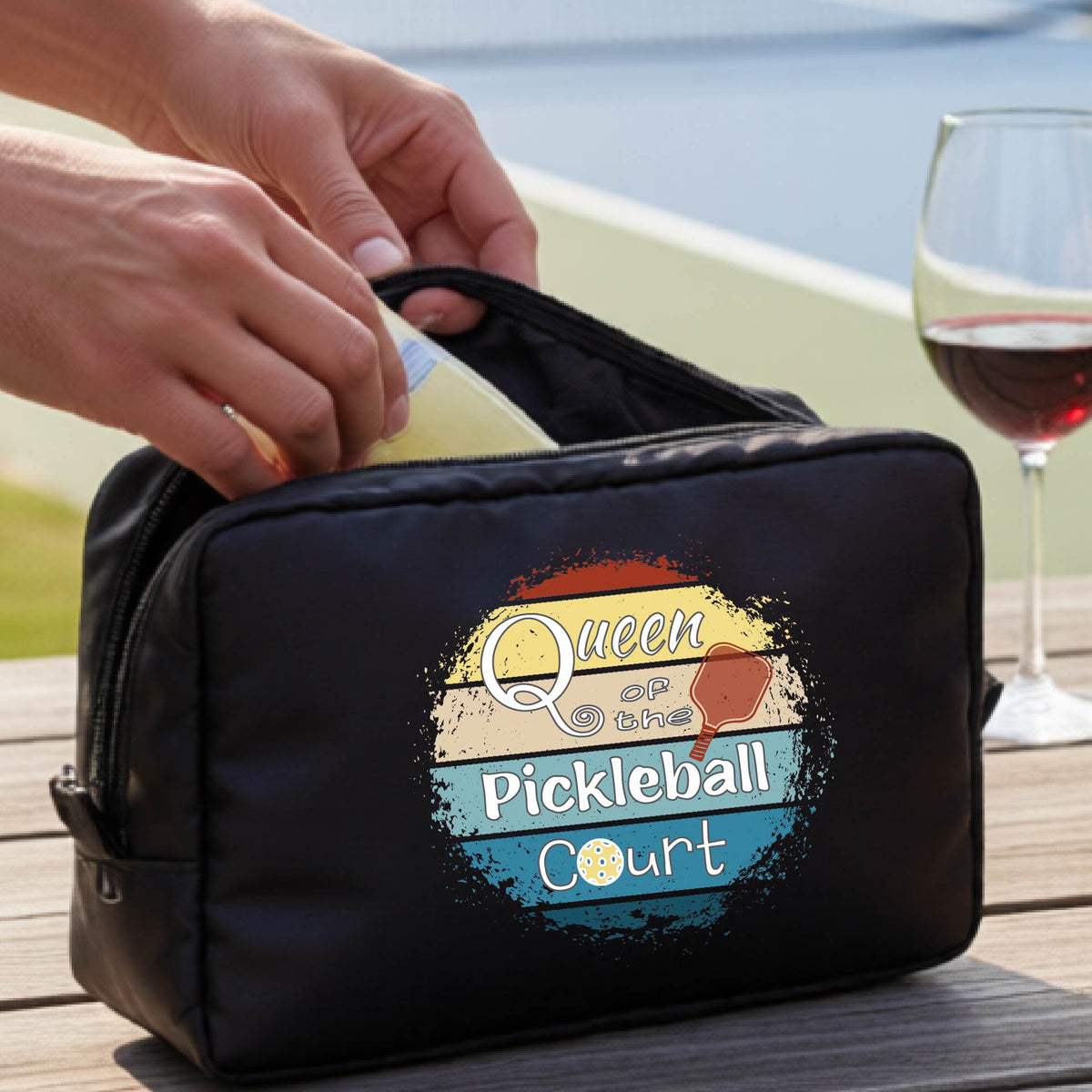 A black pickleball-themed zippered pouch with a graphic reading 'Queen of the Pickleball Court' sits on a wooden table. 