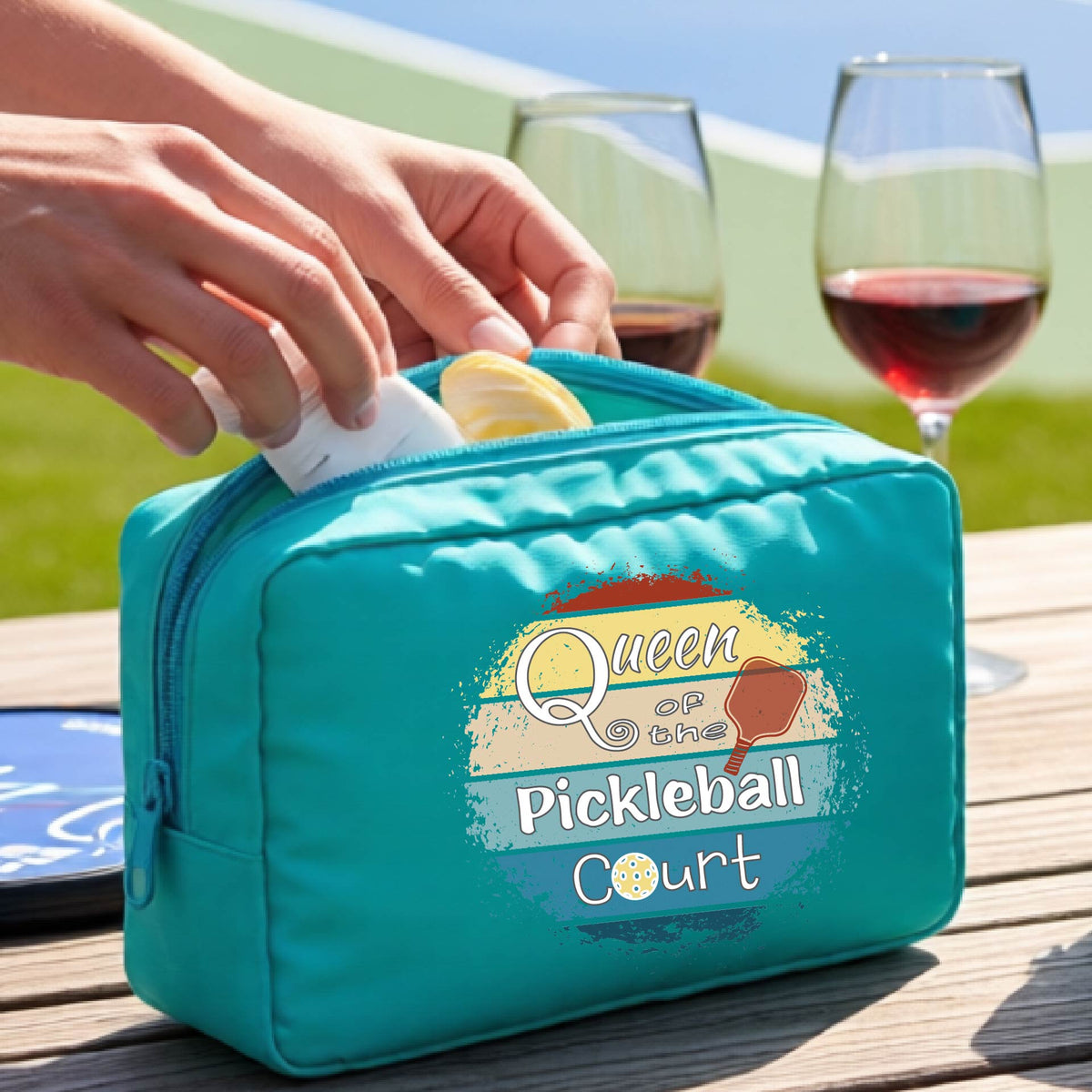 A turquoise  pickleball-themed zippered pouch with a graphic reading 'Queen of the Pickleball Court' sits on a wooden table. 