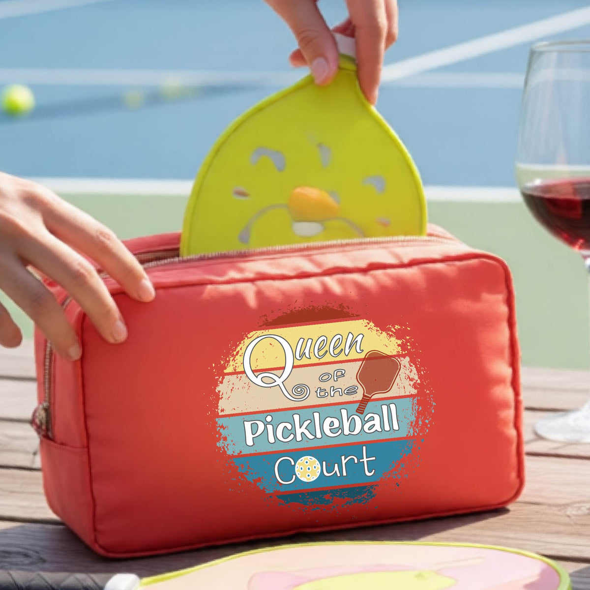 A coral-red pickleball-themed zippered pouch with a graphic reading 'Queen of the Pickleball Court' sits on a wooden table. 