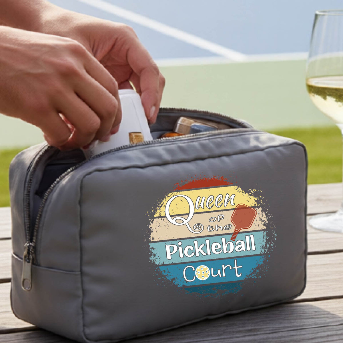 A gray pickleball-themed zippered pouch with a graphic reading 'Queen of the Pickleball Court' sits on a wooden table. 