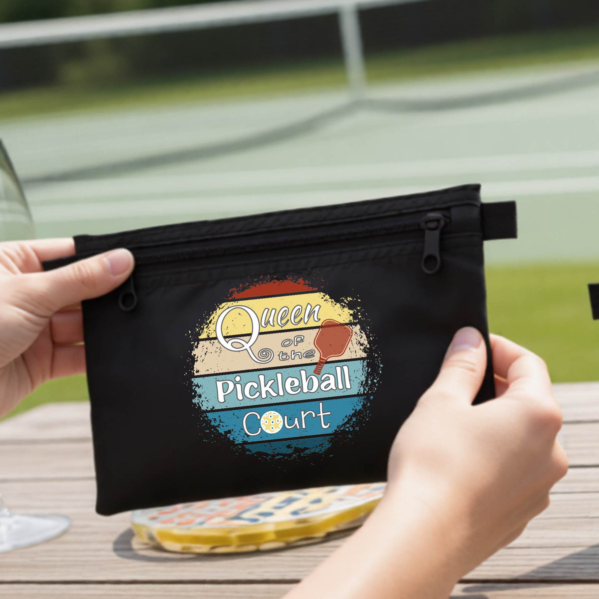 A black zippered cosmetic pouch bag with a custom 'Queen of hte Pickleball Court' graphic, being opened by hands on a wooden table.