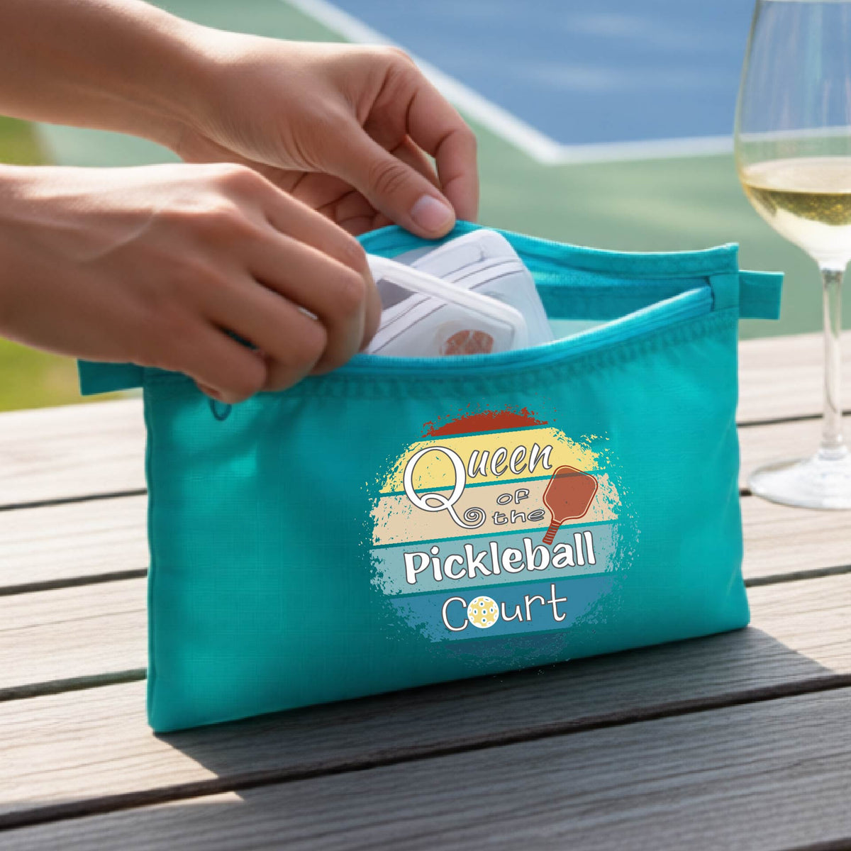 A turquoise zippered cosmetic pouch bag with a custom 'Queen of hte Pickleball Court' graphic, being opened by hands on a wooden table.
