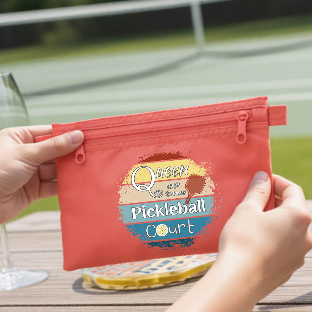 A coral colored zippered cosmetic pouch bag with a custom 'Queen of hte Pickleball Court' graphic, being opened by hands on a wooden table.