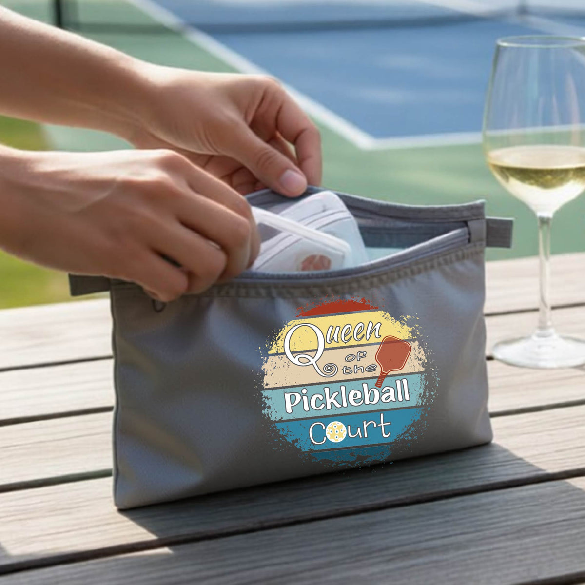 A gray zippered cosmetic pouch bag with a custom 'Queen of hte Pickleball Court' graphic, being opened by hands on a wooden table.