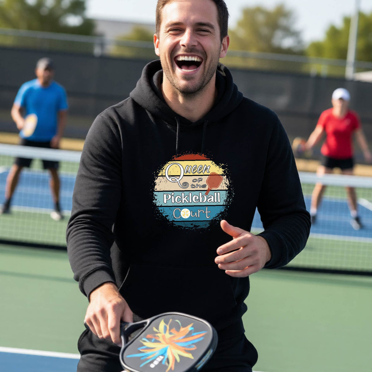 A man wearing a black hoodie with a colorful retro 'Queen of the Pickleball Court' in a retro sunset graphic stands on an outdoor pickleball court holding a paddle. Other players are visible in the background.