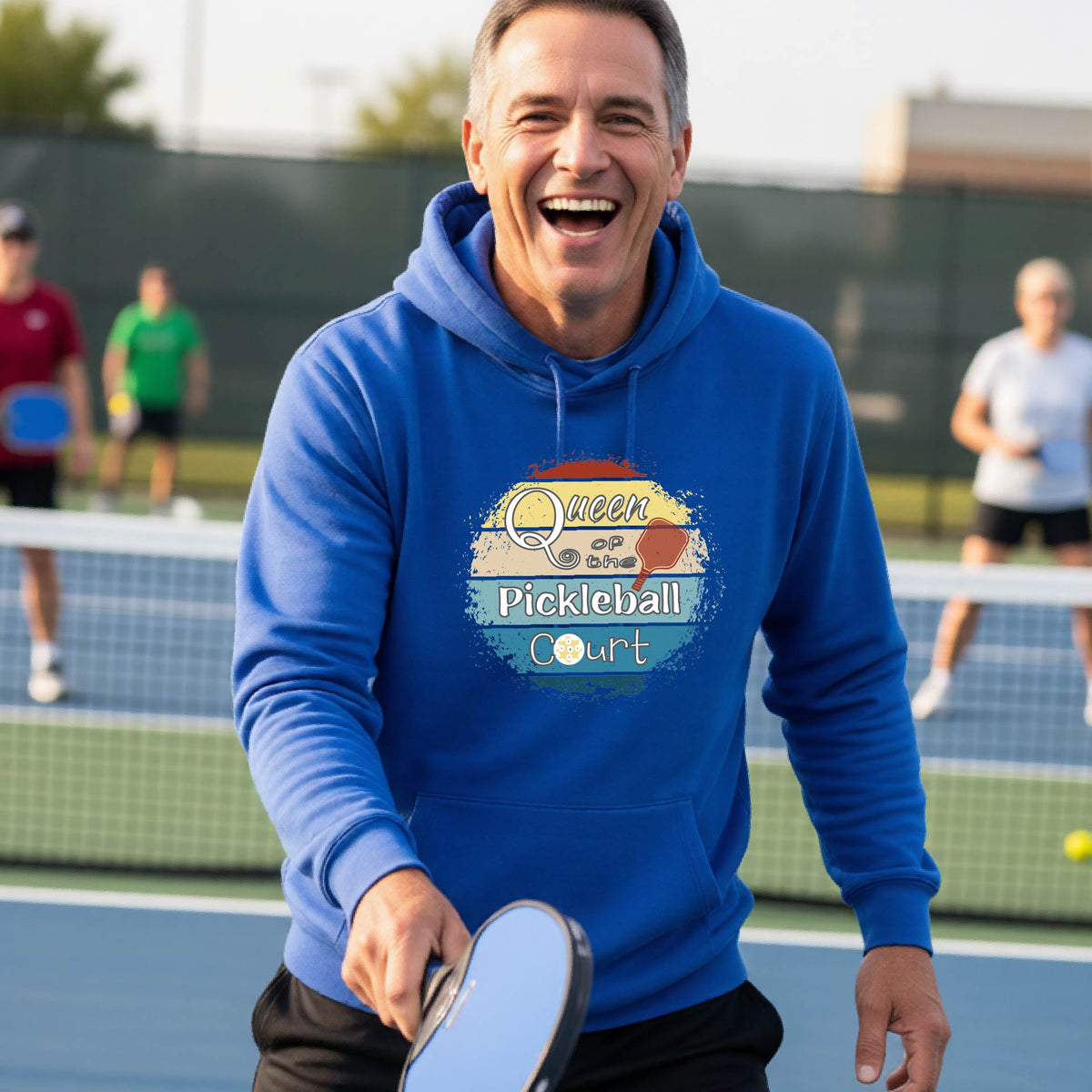 A man wearing a royal blue hoodie with a colorful retro 'Queen of the Pickleball Court' in a retro sunset graphic stands on an outdoor pickleball court holding a paddle. Other players are visible in the background.
