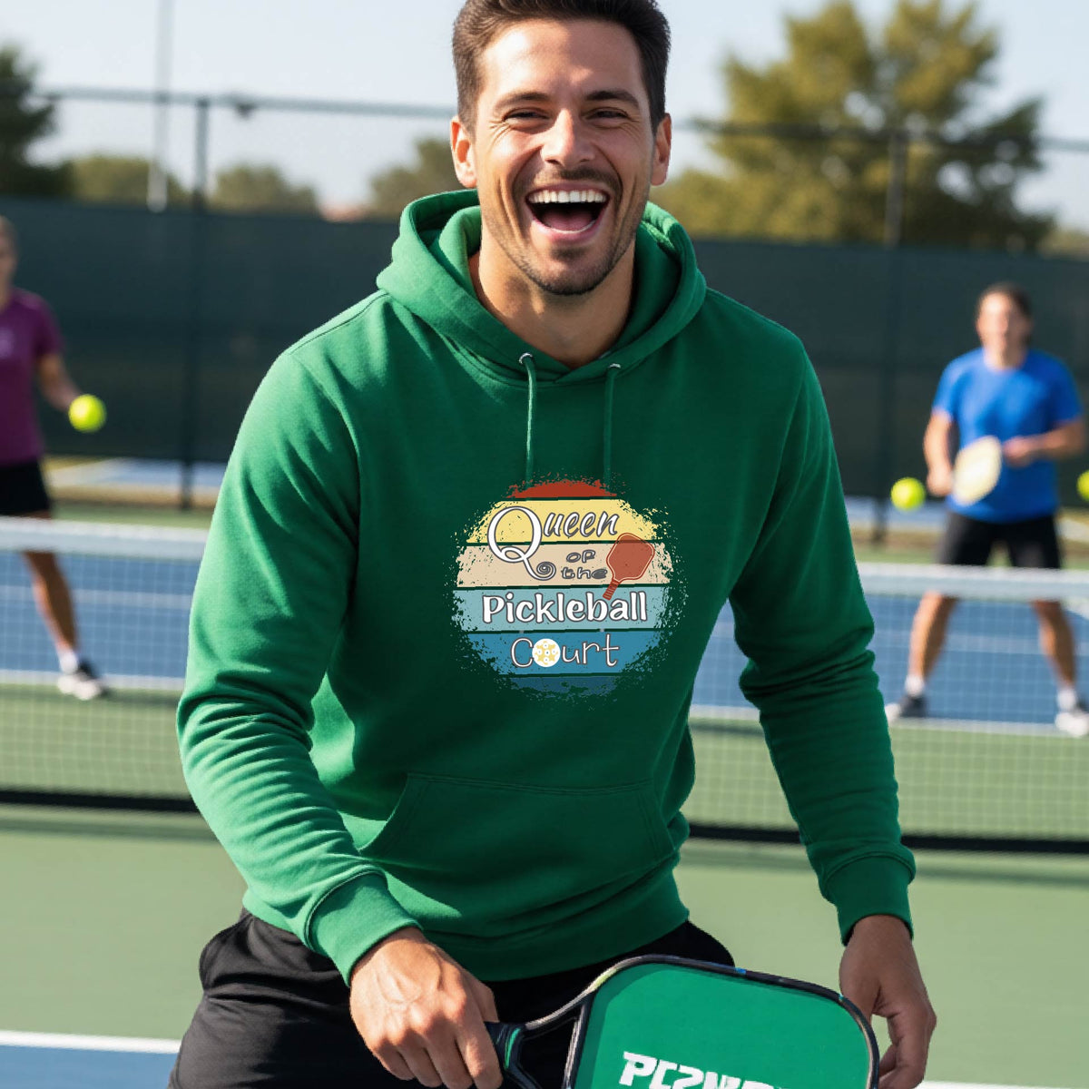 A man wearing a green hoodie with a colorful retro 'Queen of the Pickleball Court' in a retro sunset graphic stands on an outdoor pickleball court holding a paddle. Other players are visible in the background.