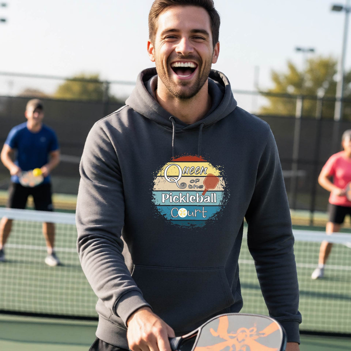 A man wearing a gray hoodie with a colorful retro 'Queen of the Pickleball Court' in a retro sunset graphic stands on an outdoor pickleball court holding a paddle. Other players are visible in the background.