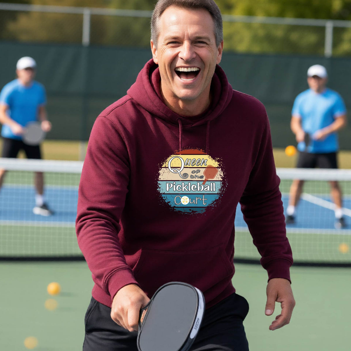 A man wearing a maroon hoodie with a colorful retro 'Queen of the Pickleball Court' in a retro sunset graphic stands on an outdoor pickleball court holding a paddle. Other players are visible in the background.