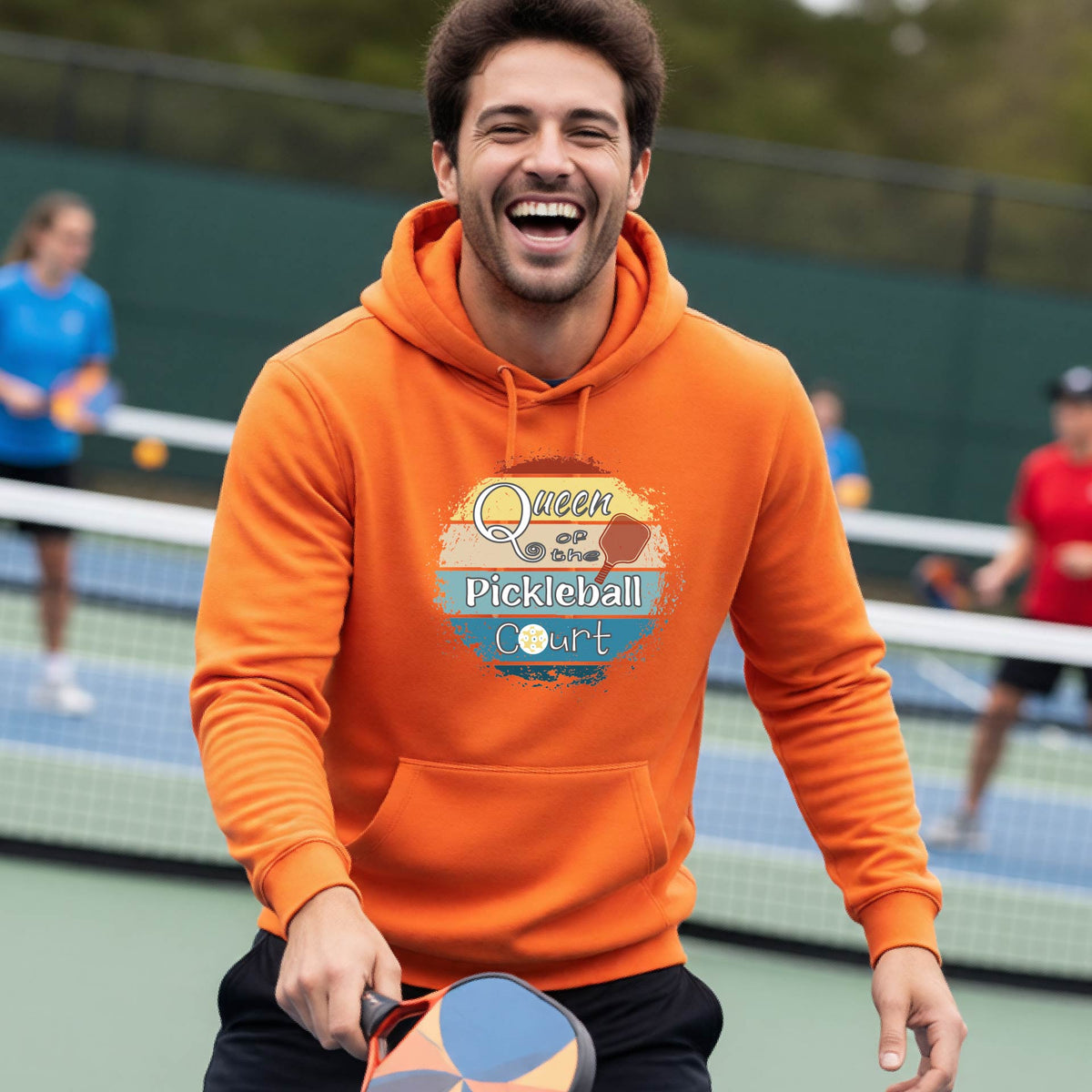 A man wearing an orange hoodie with a colorful retro 'Queen of the Pickleball Court' in a retro sunset graphic stands on an outdoor pickleball court holding a paddle. Other players are visible in the background.