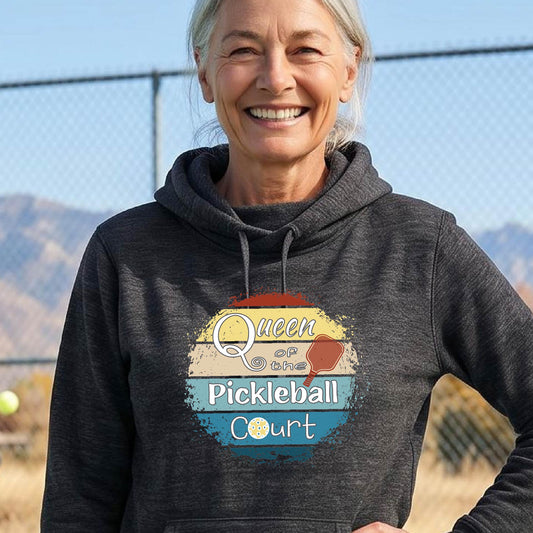A smiling older woman wearing a dark charcoal heather hoodie with a retro-style graphic that reads 'Queen of the Pickleball Court' in yellow, white, and blue, photographed outdoors on a sunny day with a chain-link fence and mountains in the background.
