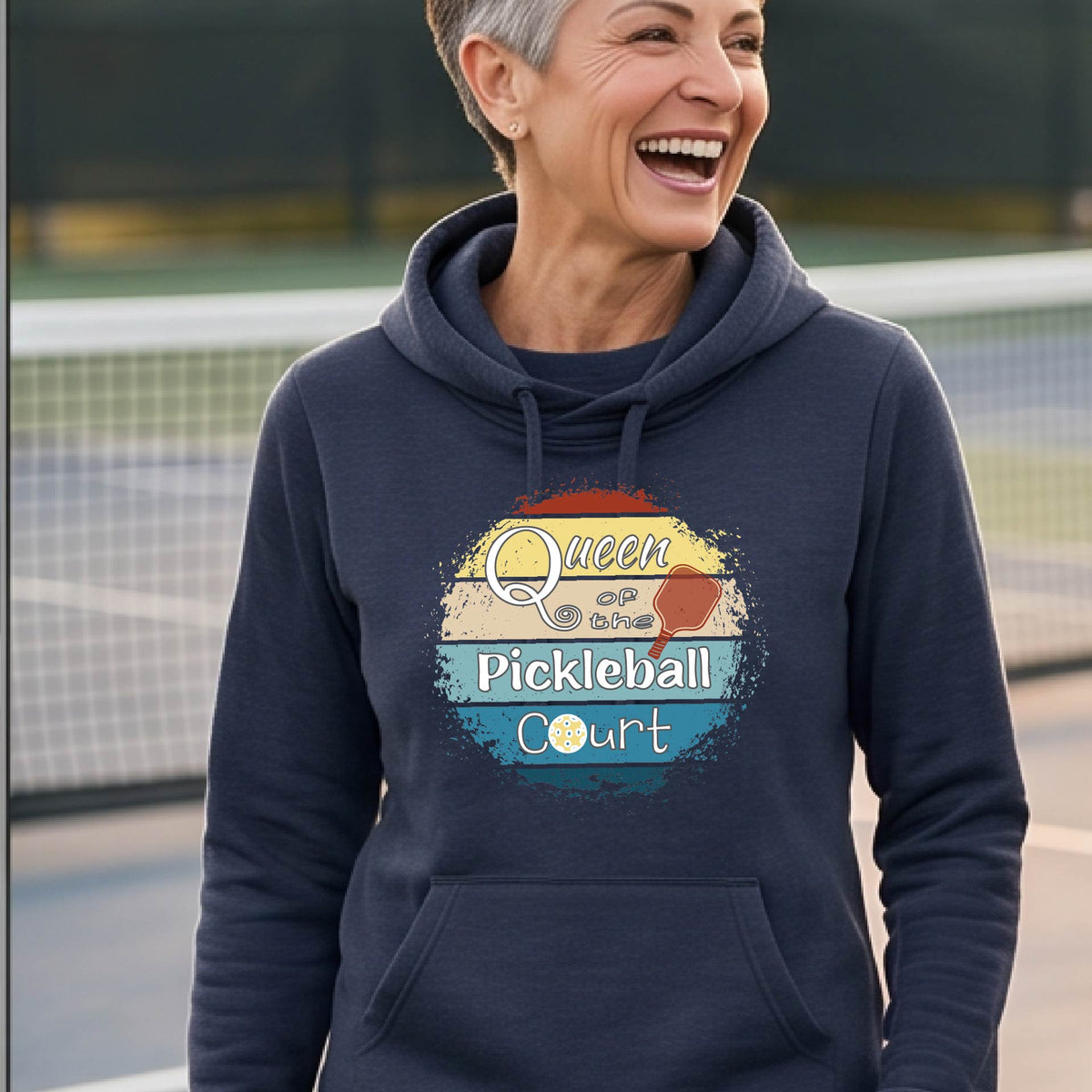 A woman wearing a navy blue hoodie with a retro-style graphic that reads 'Queen of the Pickleball Court' in yellow, white, and blue, photographed outdoors on a sunny day with a chain-link fence and mountains in the background.