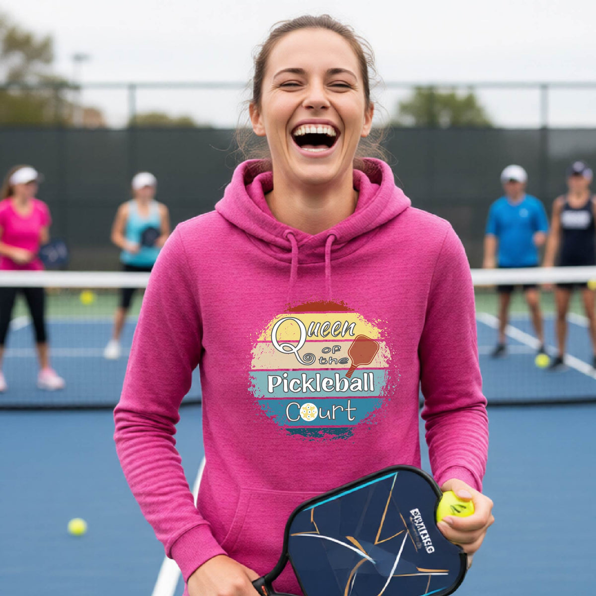 A smiling woman wearing a pink hoodie with a retro-style graphic that reads 'Queen of the Pickleball Court' in yellow, white, and blue, photographed outdoors on a sunny day with a chain-link fence and mountains in the background.