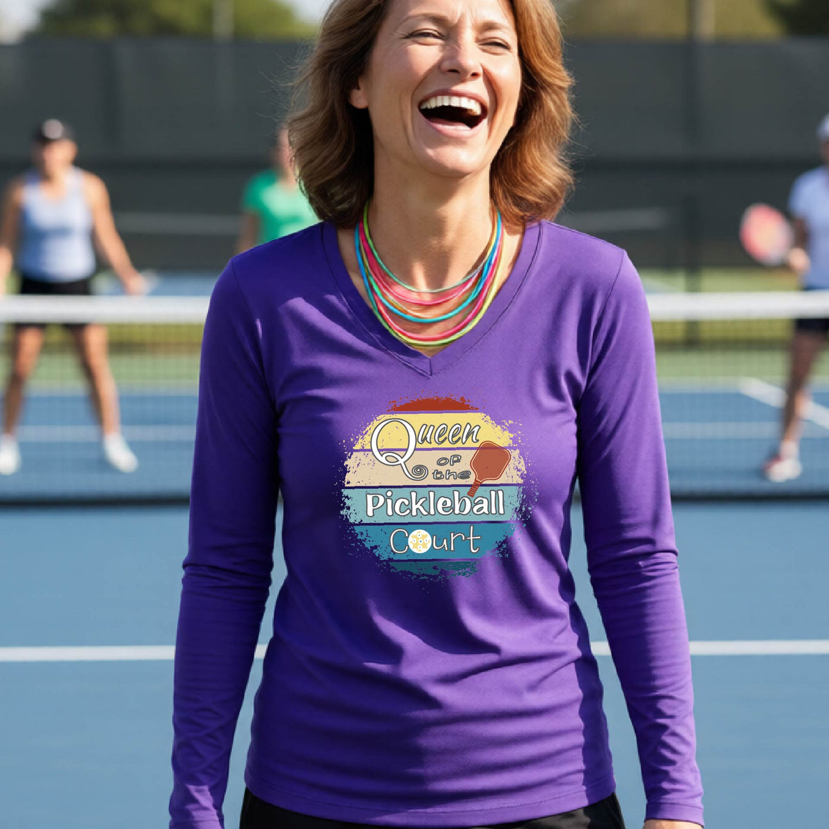 A woman laughing on an outdoor pickleball court wearing a purple long-sleeve V-neck t-shirt with a 'Queen of the Pickleball Court' with a retro sunset graphic on the chest.
