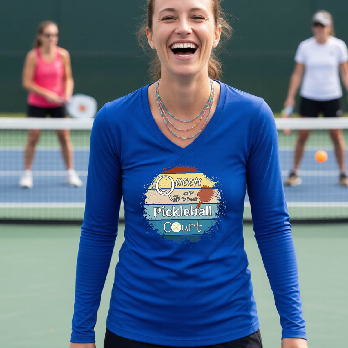 A woman laughing on an outdoor pickleball court wearing a royal blue long-sleeve V-neck t-shirt with a 'Queen of the Pickleball Court' with a retro sunset graphic on the chest.