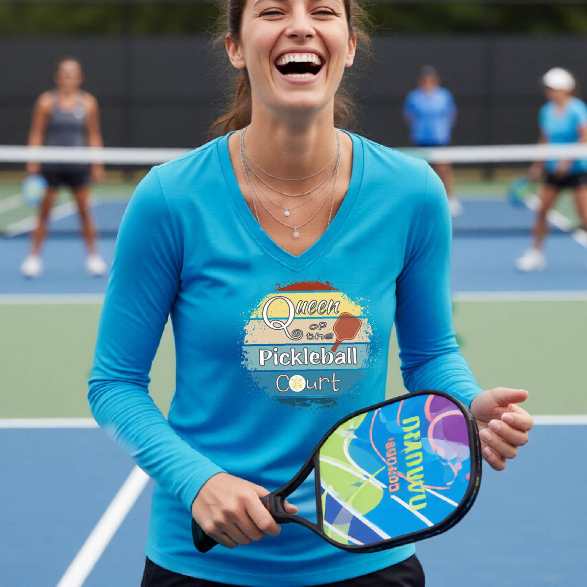 A woman laughing on an outdoor pickleball court wearing a bright blue long-sleeve V-neck t-shirt with a 'Queen of the Pickleball Court' with a retro sunset graphic on the chest.