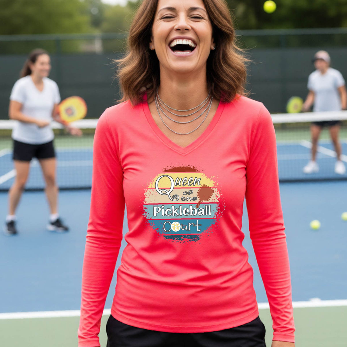 A woman laughing on an outdoor pickleball court wearing a coral pink long-sleeve V-neck t-shirt with a 'Queen of the Pickleball Court' with a retro sunset graphic on the chest.