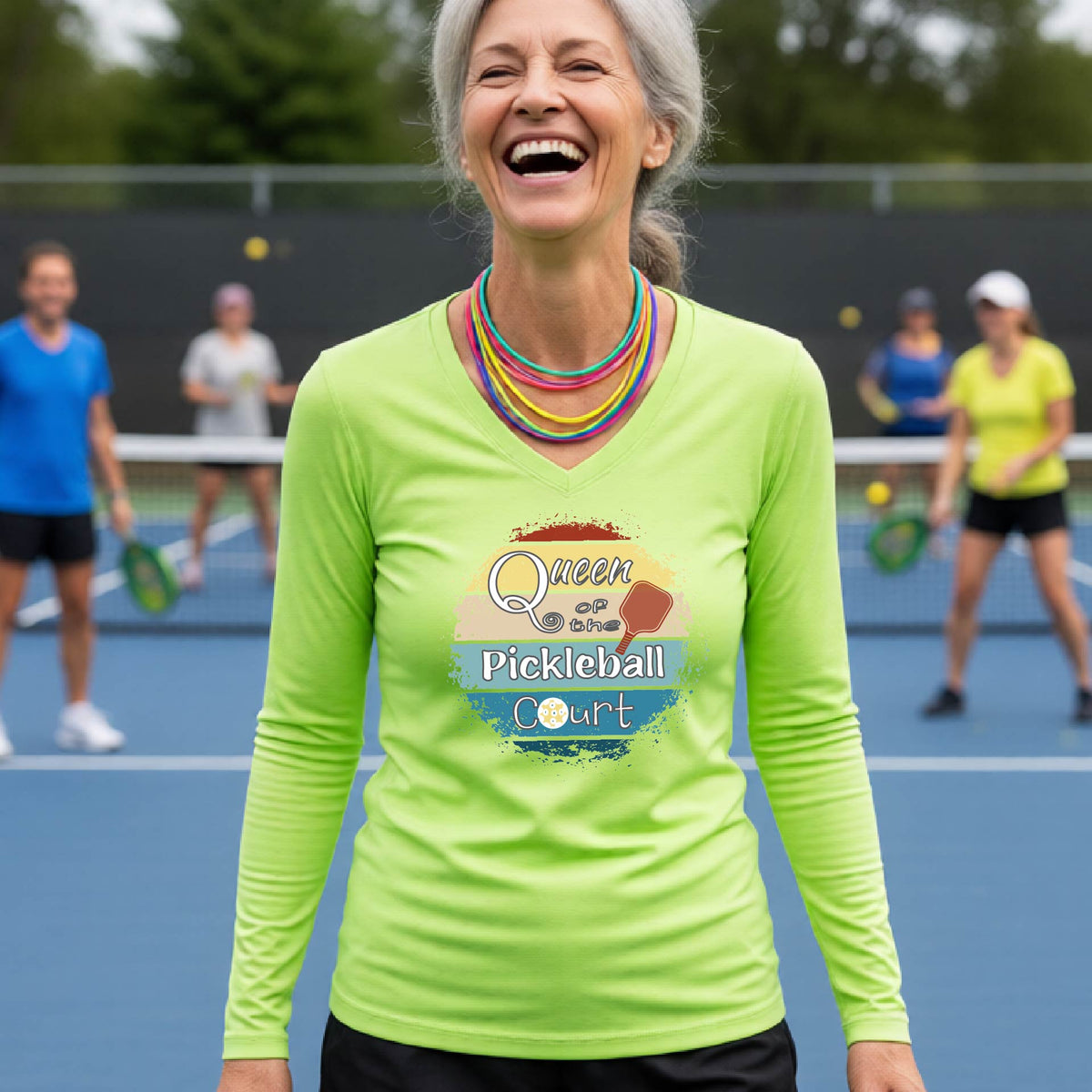 A woman laughing on an outdoor pickleball court wearing a lime green long-sleeve V-neck t-shirt with a 'Queen of the Pickleball Court' with a retro sunset graphic on the chest.