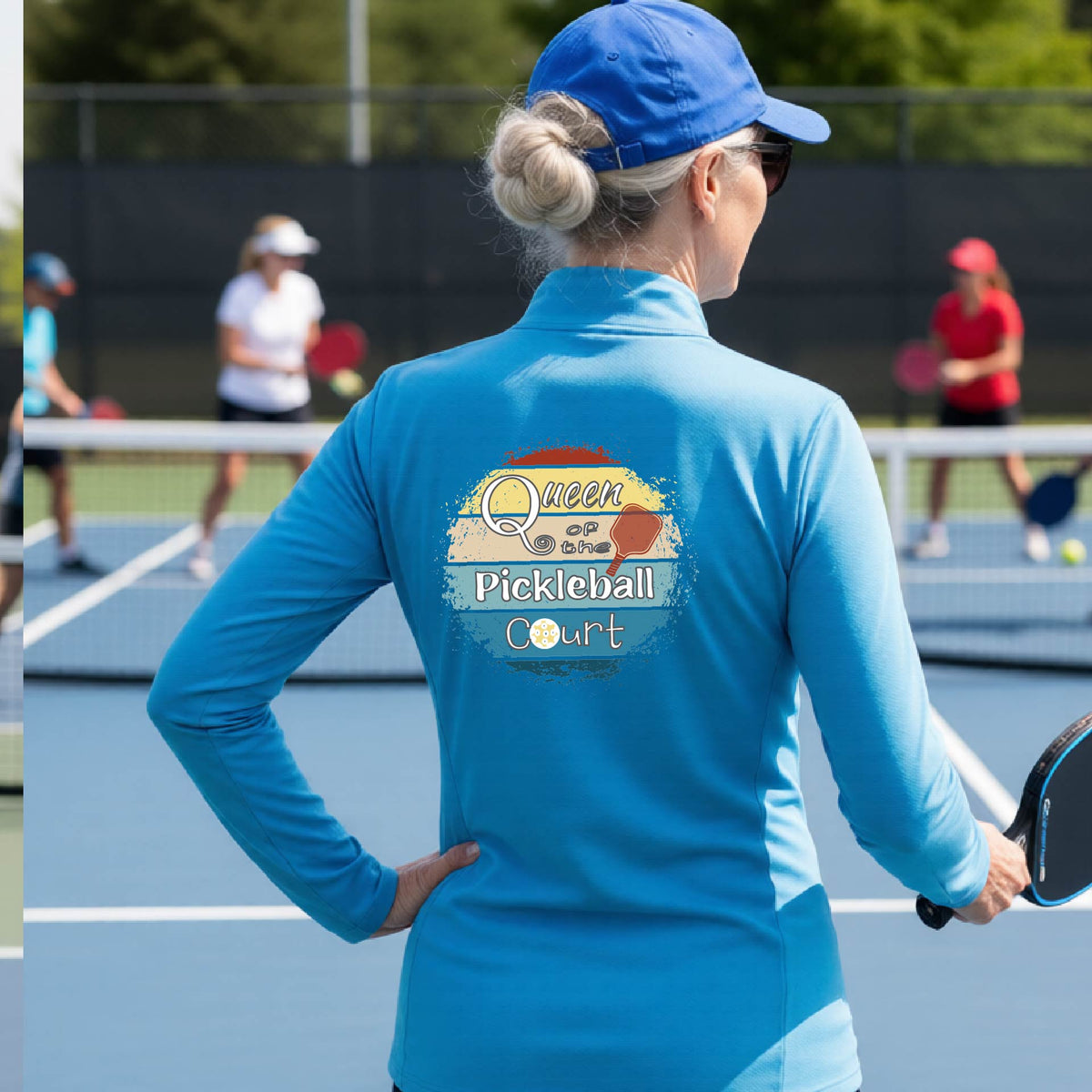 A woman on a pickleball court seen from behind, wearing a bright blue long-sleeve athletic top with a large 'Queen of hte Pickleball Court' logo printed on the back. Other players are visible in the background on the court.