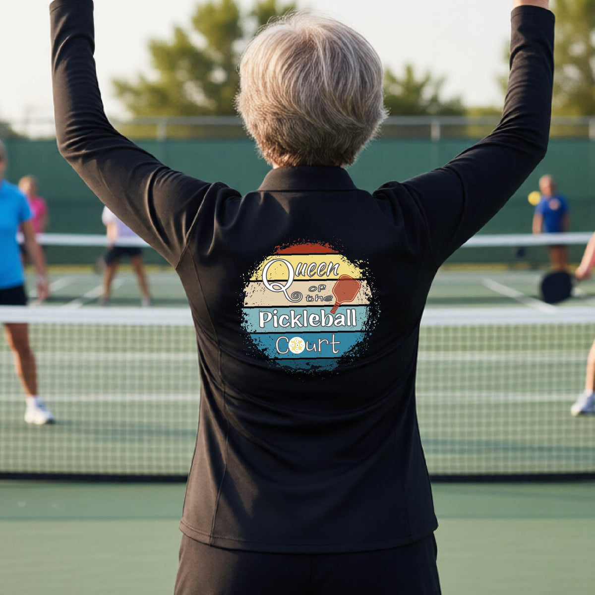 A woman on a pickleball court seen from behind, wearing a black long-sleeve athletic top with a large 'Queen of hte Pickleball Court' logo printed on the back. Other players are visible in the background on the court.
