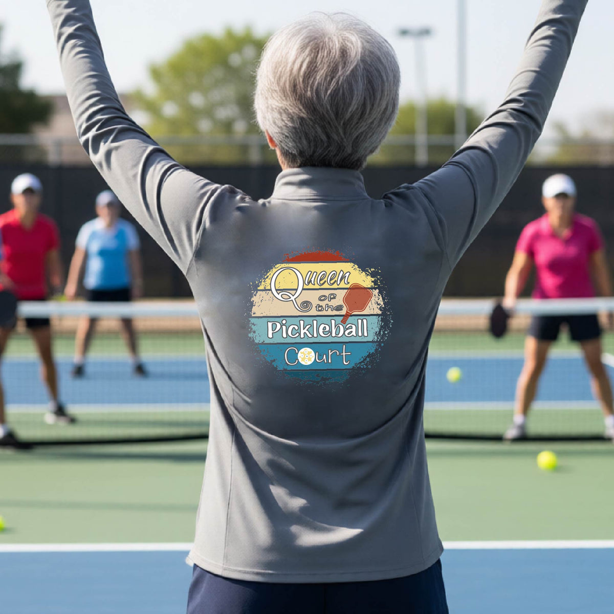 A woman on a pickleball court seen from behind, wearing a gray long-sleeve athletic top with a large 'Queen of hte Pickleball Court' logo printed on the back. Other players are visible in the background on the court.