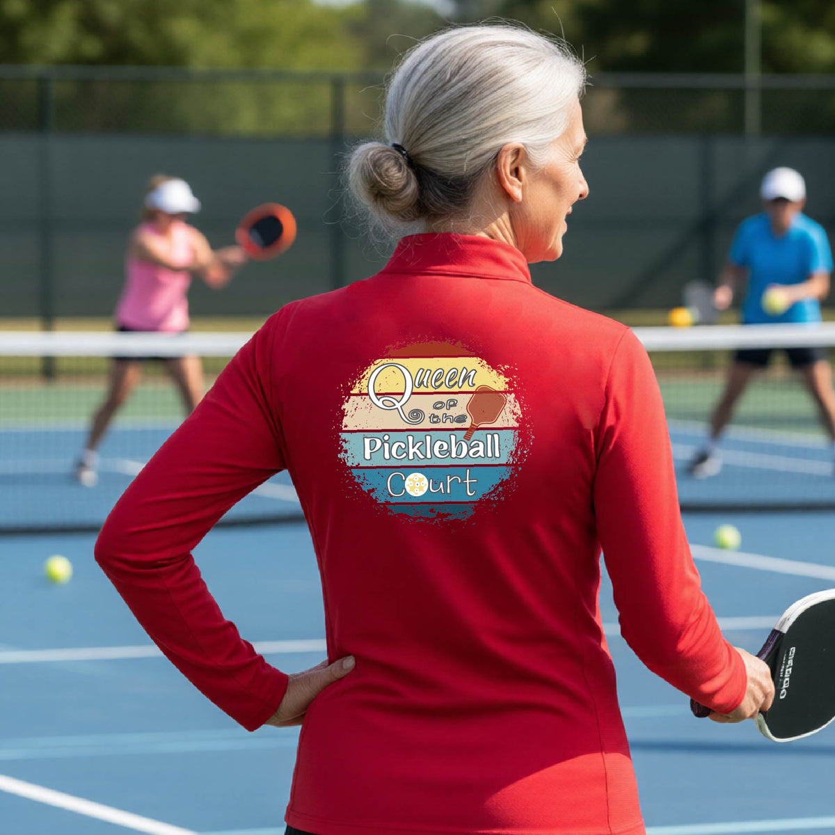 A woman on a pickleball court seen from behind, wearing a bright red long-sleeve athletic top with a large 'Queen of hte Pickleball Court' logo printed on the back. Other players are visible in the background on the court.