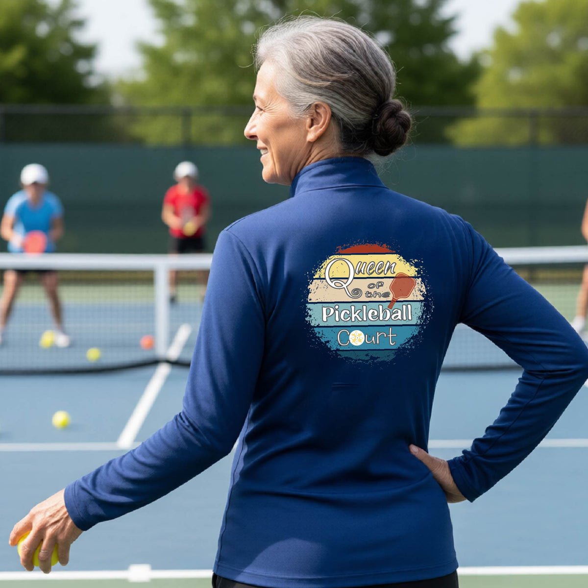 A woman on a pickleball court seen from behind, wearing a royal blue long-sleeve athletic top with a large 'Queen of hte Pickleball Court' logo printed on the back. Other players are visible in the background on the court.