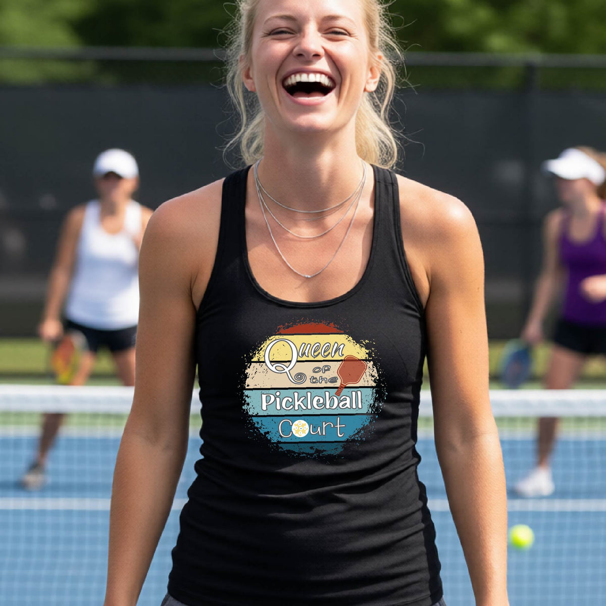 A woman laughing on a pickleball court, wearing a black racerback tank top with a retro-style graphic that reads 'Queen of the Pickleball Court' in a yellow, orange, and blue retro sunset design.