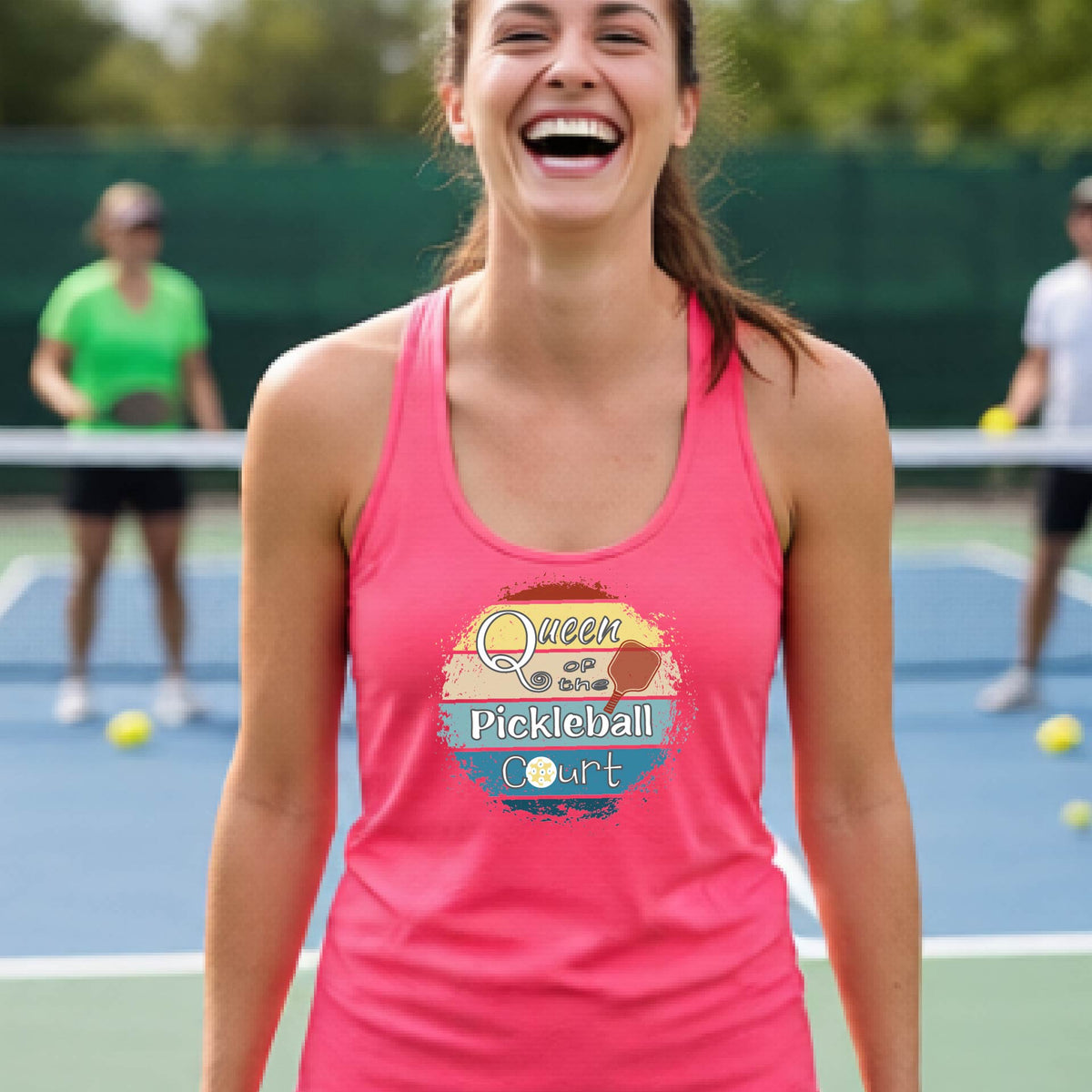 A woman laughing on a pickleball court, wearing a coral pink racerback tank top with a retro-style graphic that reads 'Queen of the Pickleball Court' in a yellow, orange, and blue retro sunset design.