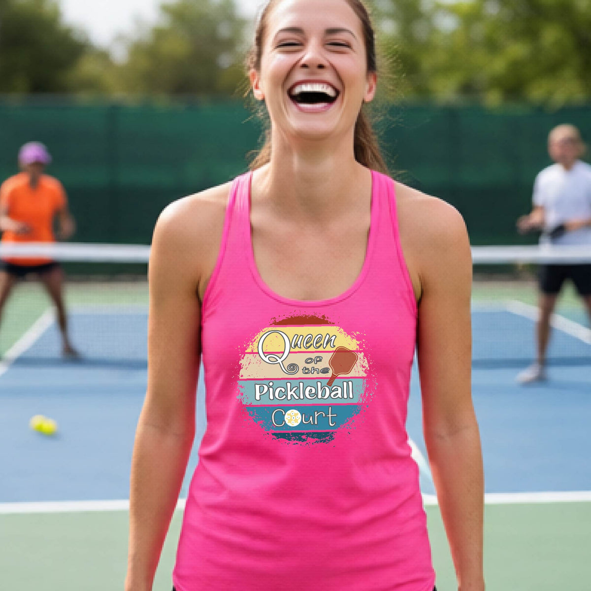 A woman laughing on a pickleball court, wearing a pink racerback tank top with a retro-style graphic that reads 'Queen of the Pickleball Court' in a yellow, orange, and blue retro sunset design.