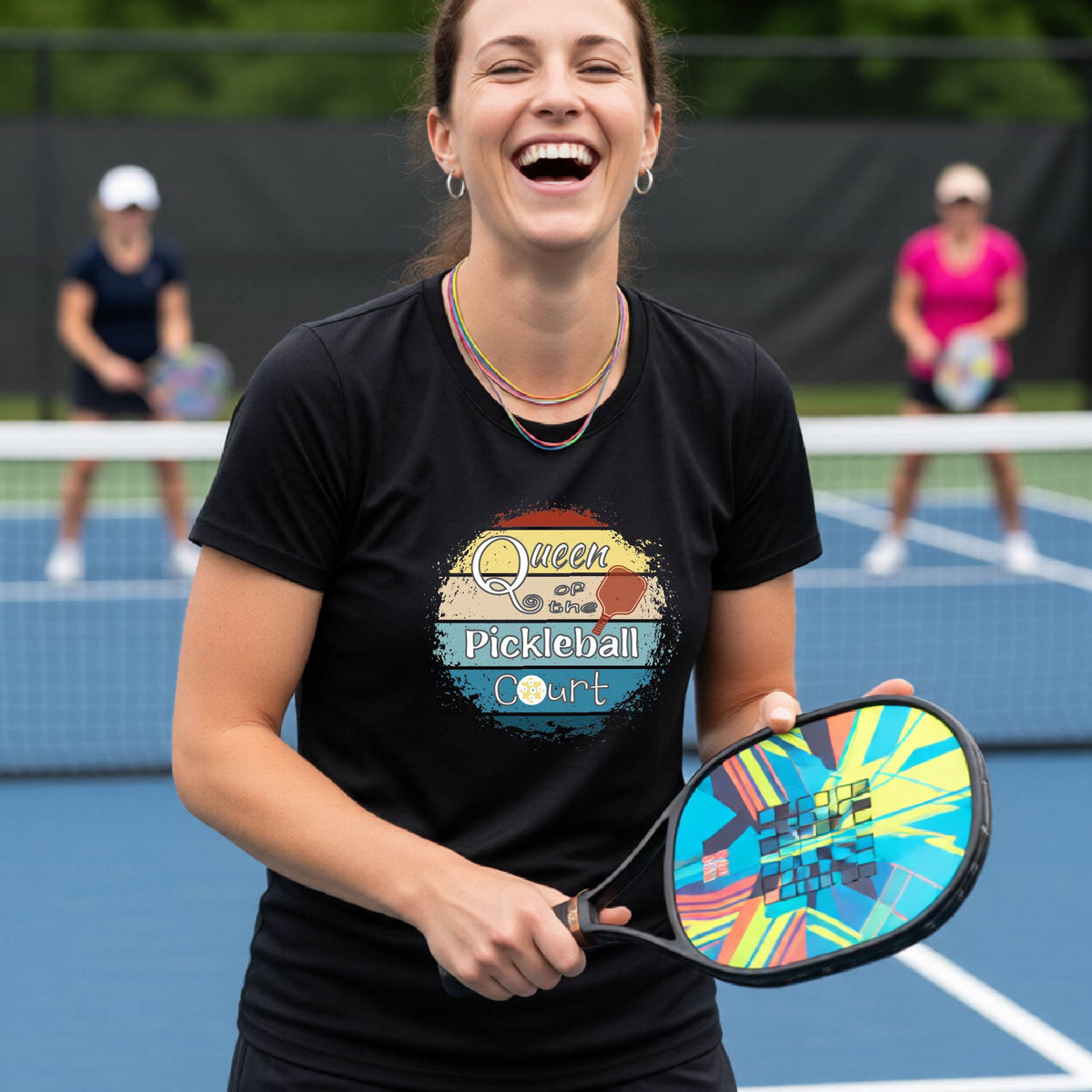 A woman on a pickleball court wearing a black t-shirt with a 'Queen of the Pickleball Court' retro sunset graphic, holding a paddle and smiling. Other players are visible in the background on the court.