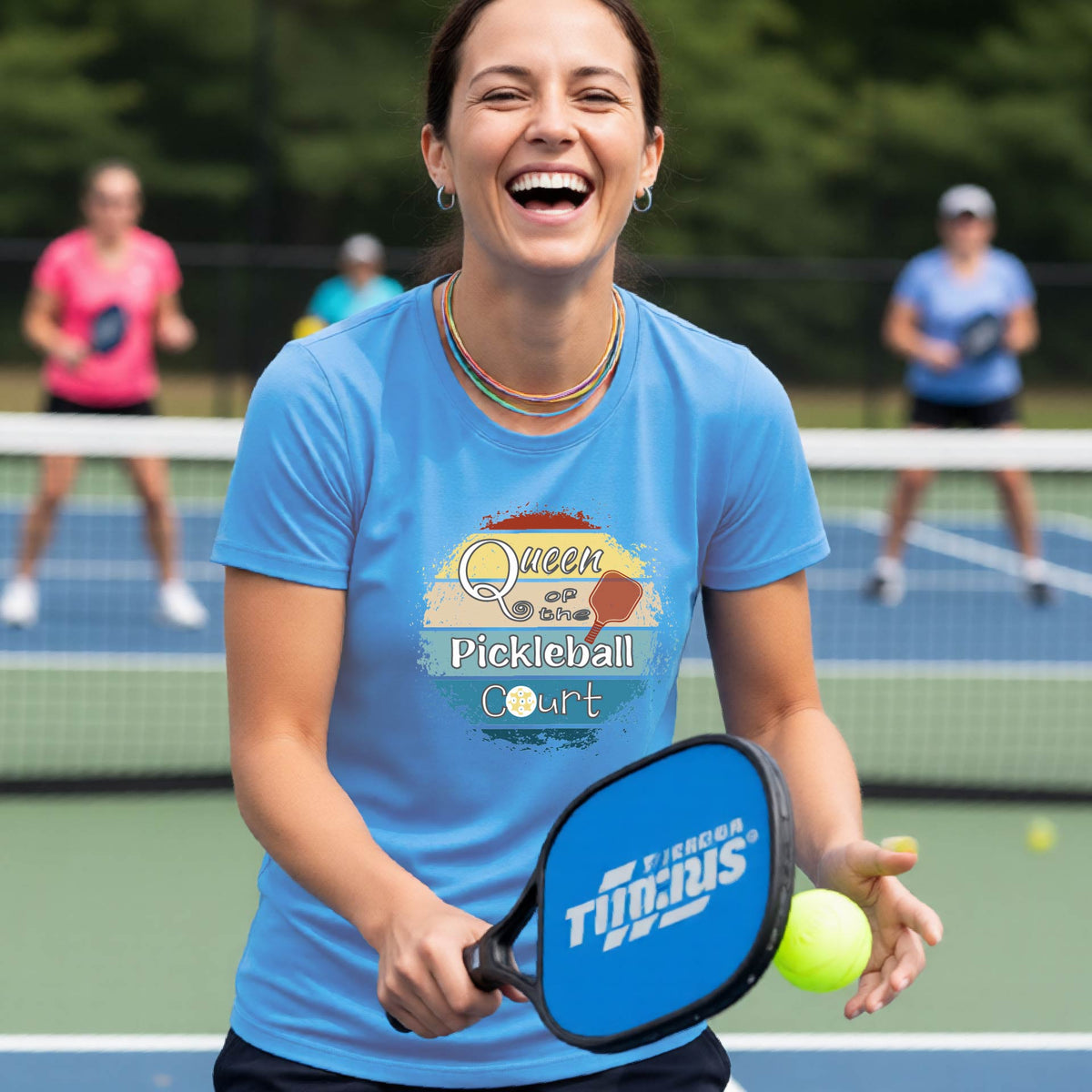 A woman on a pickleball court wearing a blue t-shirt with a 'Queen of the Pickleball Court' retro sunset graphic, holding a paddle and smiling. Other players are visible in the background on the court.