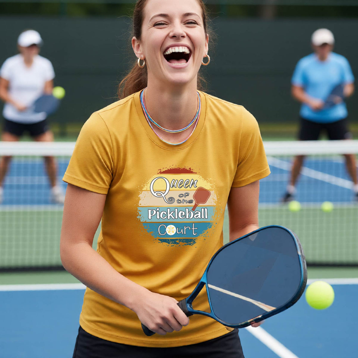 A woman on a pickleball court wearing a mustard yellow gold t-shirt with a 'Queen of the Pickleball Court' retro sunset graphic, holding a paddle and smiling. Other players are visible in the background on the court.