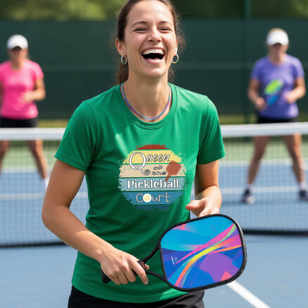 A woman on a pickleball court wearing a green t-shirt with a 'Queen of the Pickleball Court' retro sunset graphic, holding a paddle and smiling. Other players are visible in the background on the court.