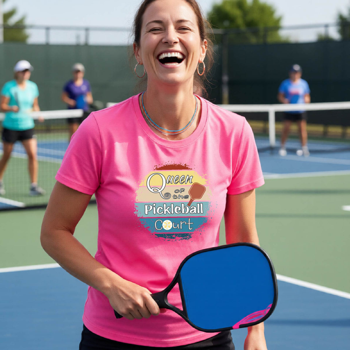 A woman on a pickleball court wearing a pink t-shirt with a 'Queen of the Pickleball Court' retro sunset graphic, holding a paddle and smiling. Other players are visible in the background on the court.